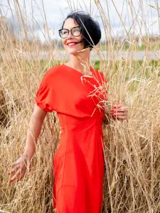Een vrouw in een rode jurk staat tussen droge grassprietjes en glimlacht met gesloten ogen.
