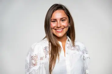 Een glimlachende vrouw met lang bruin haar, dragen een witte blouse met decoratieve openingen op de schouders, staand voor een effen lichte achtergrond.