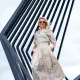 Een vrouw in een beige, bloemendekleding staat voor een moderne, geometrische metalen structuur, glimlacht en draagt een zonnebril.