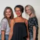 Drie vrouwen glimlachen naar de camera, staand dicht bij elkaar, tegen een witte achtergrond.